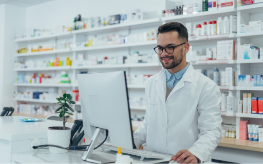 pharmacist stands in front of a wall of medication looking at a computer