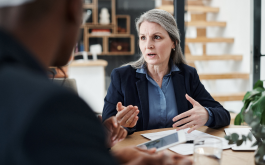 mature woman in suit sitting at desk talking to someone out of frame
